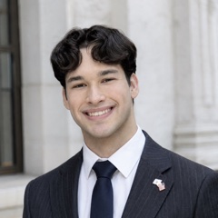 Portrait of Emmanuel Colmenares smiling in a navy pinstripe suit and navy tie, with an American flag pin on his lapel, in front of a white marble building.