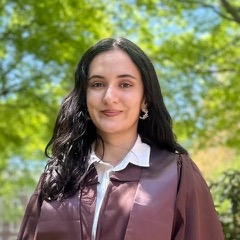 Portrait of Yosumin Qurbonbekova in a Brown University graduation gown, on a tree-lined campus path.